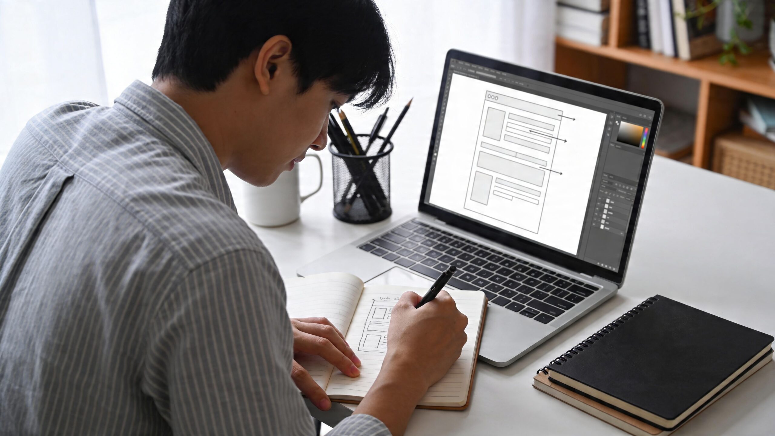 A focused website designer sketches interface wireframes in a notebook while working on a laptop at his desk.